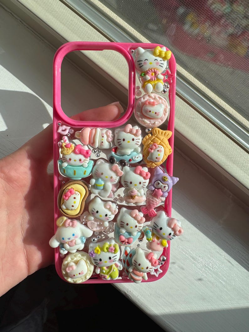May include: A pink phone case decorated with multiple Hello Kitty character charms. The charms feature different poses and outfits, with pastel colours and glitter. The case is held in a hand near a window.
