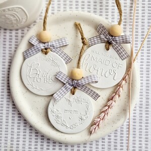 May include: Three white ceramic ornaments with twine and a gray and white gingham ribbon. The ornaments say "Bride to Be", "Will you be my Maid of Honor", and "Mum".