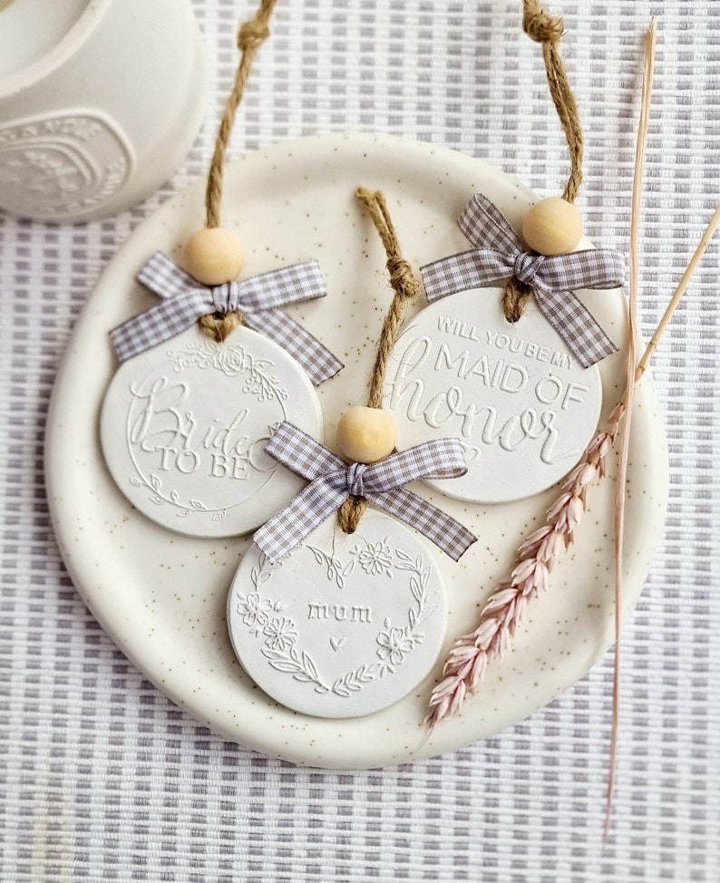 May include: Three white ceramic ornaments with twine and a gray and white gingham ribbon. The ornaments say "Bride to Be", "Will you be my Maid of Honor", and "Mum".