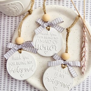 May include: Three white ceramic ornaments with twine and a wooden bead. Each ornament has a different message: "Thank You For Being My Maid of Honor", "Thank You For Being My Bridesmaid", and "Will You Be My Bridesmaid".