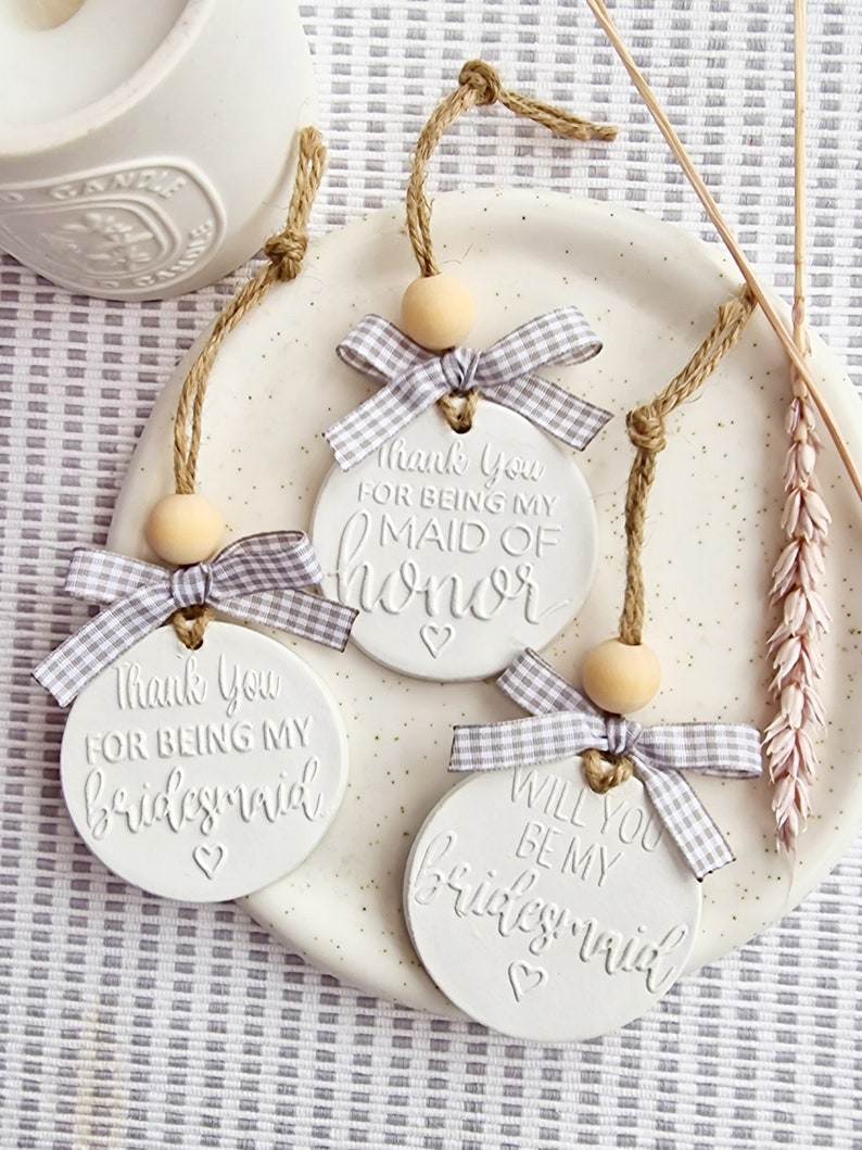 May include: Three white ceramic ornaments with twine and a wooden bead. Each ornament has a different message: "Thank You For Being My Maid of Honor", "Thank You For Being My Bridesmaid", and "Will You Be My Bridesmaid".