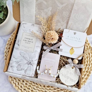 May include: A gift box filled with a variety of items, including a cookie crumble candle, a pair of white hoop earrings, a necklace with a gold leaf charm, and a ceramic ornament that says "Will you be my bridesmaid?" The box is lined with brown paper and has a white floral pattern.