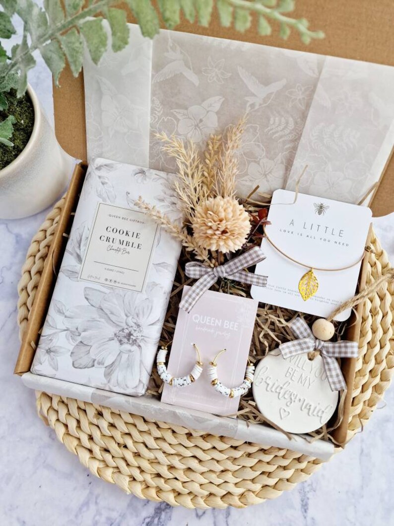 May include: A gift box filled with a variety of items, including a cookie crumble candle, a pair of white hoop earrings, a necklace with a gold leaf charm, and a ceramic ornament that says "Will you be my bridesmaid?" The box is lined with brown paper and has a white floral pattern.
