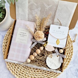 May include: A gift box filled with items for a bridesmaid. The box contains a pink and white cookie crumble candle, a wooden bead necklace, a keychain with a pink leaf charm, a small card with the text "A little love is all you need", and a white ceramic ornament with the text "Will you be my bridesmaid?"