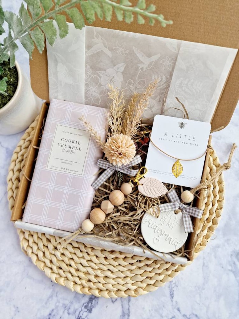 May include: A gift box filled with items for a bridesmaid. The box contains a pink and white cookie crumble candle, a wooden bead necklace, a keychain with a pink leaf charm, a small card with the text "A little love is all you need", and a white ceramic ornament with the text "Will you be my bridesmaid?"