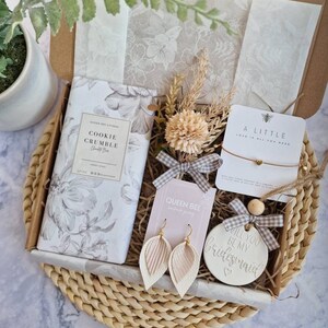 May include: A gift box filled with a variety of items, including a cookie crumble scented bar, a pair of earrings, a small ceramic ornament that says "Will you be my bridesmaid?" and a small card that says "A little love is all you need".
