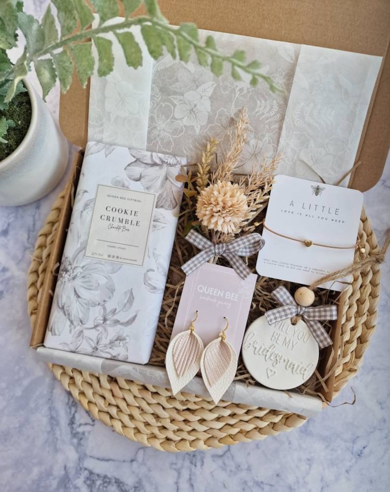 May include: A gift box filled with a variety of items, including a cookie crumble scented bar, a pair of earrings, a small ceramic ornament that says "Will you be my bridesmaid?" and a small card that says "A little love is all you need".