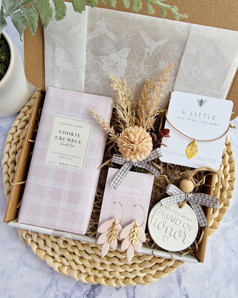 May include: A gift box filled with items for a maid of honor. The box includes a cookie crumble bar, a pair of gold and pink earrings, a small wooden sign that says "Will you be my Maid of Honor", and a small card that says "A Little Love is All You Need".
