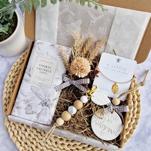 May include: A gift box filled with items for a bridesmaid. The box contains a candle, a small wooden bead necklace, a ceramic ornament that says "All you be my bridesmaid", a small bag of bath salts, and a necklace with a gold leaf charm. The box is decorated with a brown and white gingham ribbon.