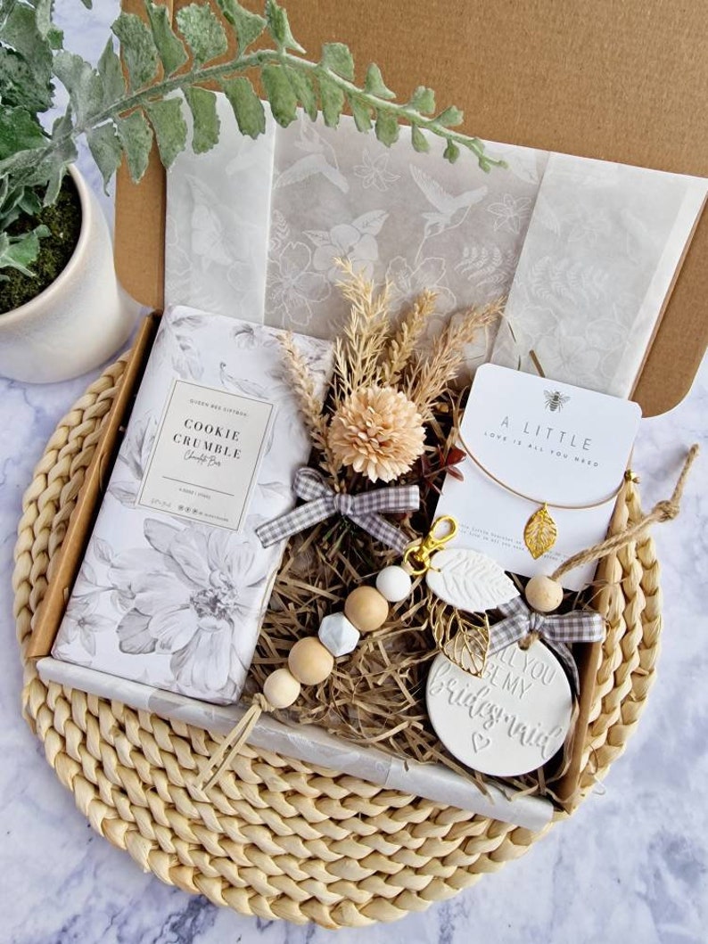 May include: A gift box filled with items for a bridesmaid. The box contains a candle, a small wooden bead necklace, a ceramic ornament that says "All you be my bridesmaid", a small bag of bath salts, and a necklace with a gold leaf charm. The box is decorated with a brown and white gingham ribbon.