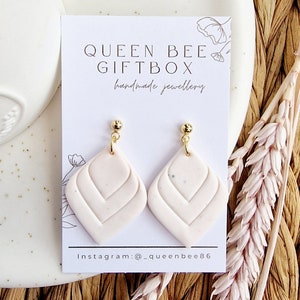 May include: Light pink, handmade earrings with a geometric pattern, presented on a white card. The card reads "QUEEN BEE GIFTBOX handmade jewellery". The earrings have gold-coloured studs and a diamond-shaped design. Instagram: @_queenbee86.