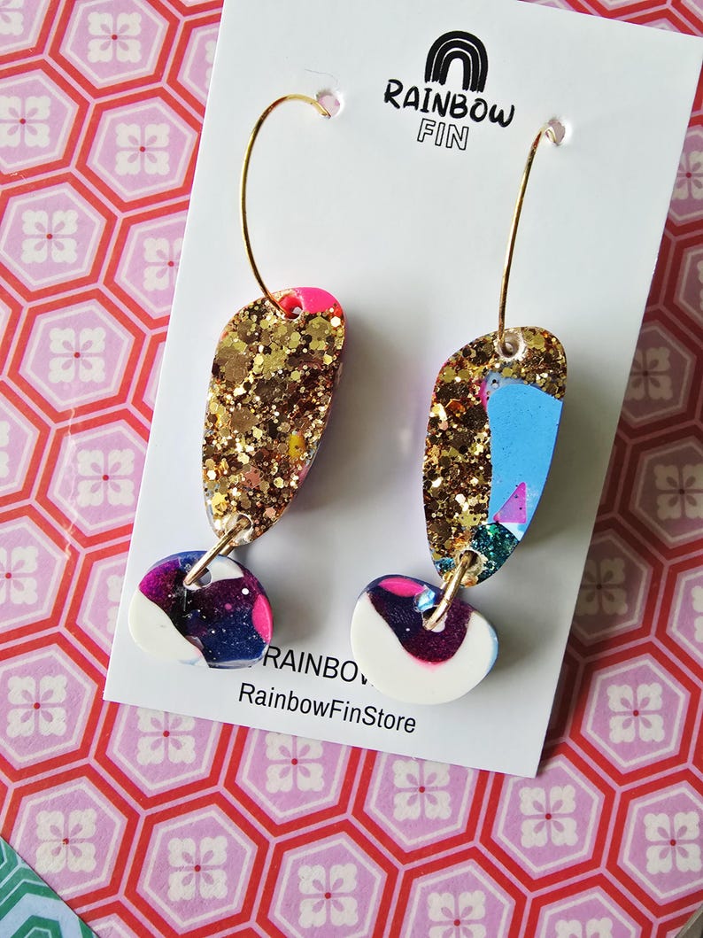 Puede incluir: Pendientes de aro dorados con un dise&ntilde;o &uacute;nico. Los pendientes presentan una parte superior con purpurina dorada, un acento rosa y una secci&oacute;n inferior con un patr&oacute;n blanco, azul y morado. Se muestran en una tarjeta blanca con el texto "RAINBOW FIN" y "RainbowFinStore".