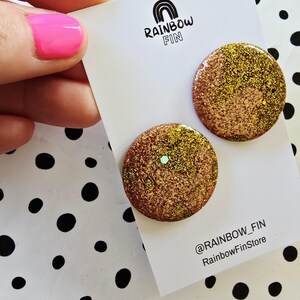 May include: Two round, glittery stud earrings in shades of gold and bronze are displayed on a white card. The card features the text "RAINBOW FIN" and the shop's social media handle. The background is a white surface with black polka dots.