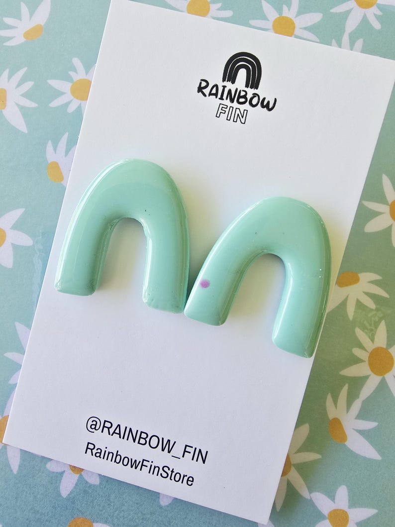 Op de afbeelding: Mintgroene, boogvormige oorbellen op een witte kaart met de tekst "RAINBOW FIN" en een regenboogafbeelding. De kaart bevat ook de tekst "@RAINBOW_FIN RainbowFinStore". De achtergrond heeft een madeliefjespatroon.