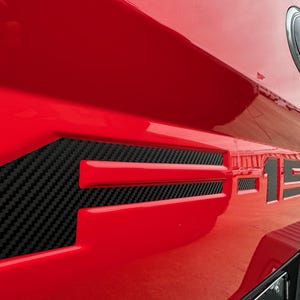 May include: Close-up of a red vehicle's tailgate with the F-150 logo. The letter "F" is outlined in red with a carbon fiber insert. The "150" is in black with a carbon fiber pattern.