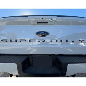May include: Rear view of a white Ford Super Duty truck. The Ford logo is centered above the black "SUPER DUTY" lettering. The tailgate features a black bumper and license plate area, with a camera and lock mechanism visible.