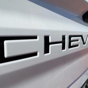 May include: A white truck tailgate featuring the word CHEVROLET embossed in large, bold, black capital letters. The lettering is recessed into the white metal surface, creating a high-contrast visual effect across the rear of the vehicle.