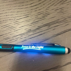 May include: A teal blue pen with a black rubber grip and a stylus tip. The pen has a white inscription that reads "Jesus is the Light".