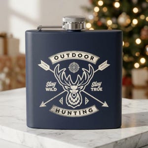 May include: A navy blue metal flask with a silver screw-top lid. The front features a white graphic of a deer head centred between two crossed arrows and a target symbol. The text reads OUTDOOR HUNTING, Stay WILD, and Stay TRUE.