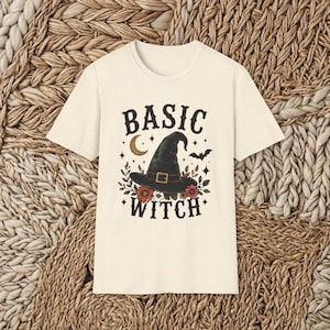 May include: A cream-colored short-sleeve t-shirt featuring a graphic print of a black witch hat, a crescent moon, a bat, and colorful flowers. The text "BASIC WITCH" is printed in a bold, dark font above and below the central illustration.