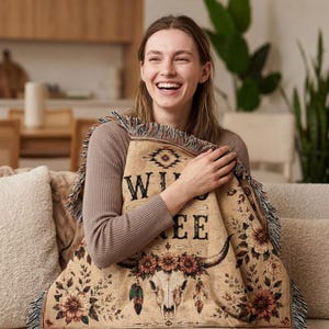 Peut inclure: Un plaid tissé sur fond beige avec un motif central représentant un crâne de bœuf orné de fleurs, de plumes et de motifs géométriques. Le texte "WILD FREE" est inscrit au-dessus du crâne et les bords présentent une frange décorative.
