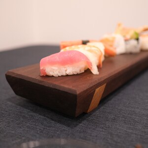 May include: A wooden sushi serving board with a variety of sushi pieces, including tuna, salmon, and shrimp. The board is dark brown with a light brown accent.