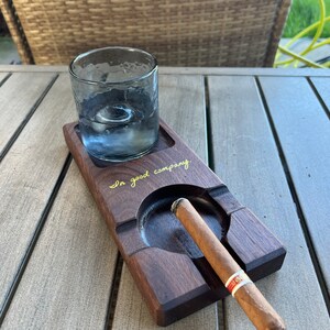 May include: A wooden tray with a glass and a cigar resting in the designated slots. The tray is engraved with the words "In good company".