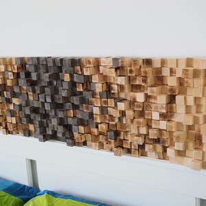 May include: Wooden headboard with a gradient design of small, square blocks. The blocks transition from light brown to dark brown, creating a textured, three-dimensional effect. The headboard is mounted above a white bed frame.