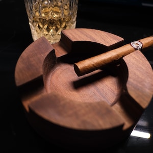 May include: A wooden cigar ashtray with a brown cigar resting on the edge. The ashtray has a circular shape with four sections.