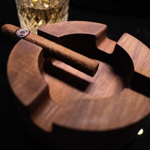 May include: A wooden cigar ashtray with a brown cigar resting on the edge. The ashtray has a deep well for ashes and a raised lip to prevent spills.