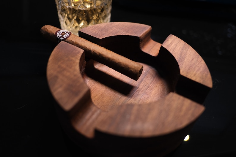 May include: A wooden cigar ashtray with a brown cigar resting on the edge. The ashtray has a deep well for ashes and a raised lip to prevent spills.