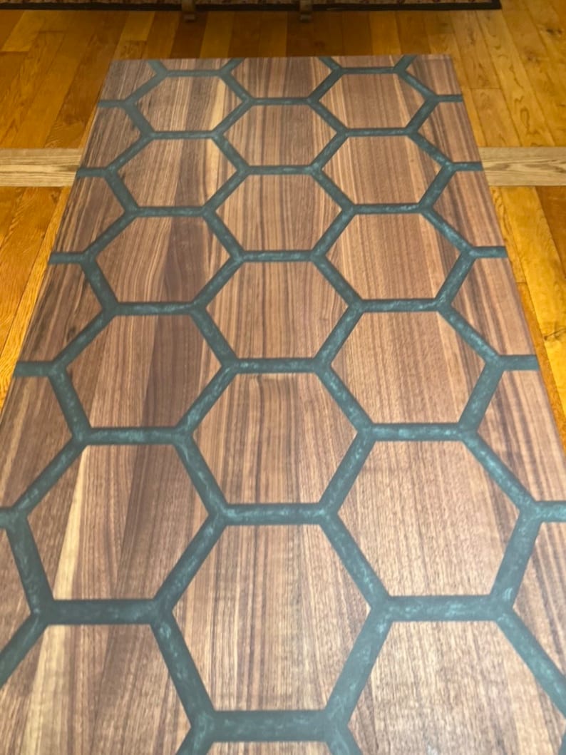 May include: A rectangular wooden table with a honeycomb design. The table's surface features a natural wood grain pattern, with dark gray epoxy resin filling the hexagonal cells. The table is a medium brown color.