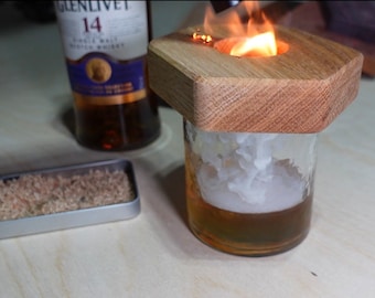 May include: A wooden coaster with a small hole in the center, holding a glass of amber liquid with a white foam on top. The coaster is on a wooden surface and there is a flame burning in the hole. A bottle of Glenlivet 14 year old single malt scotch whisky is in the background.