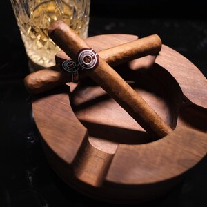 May include: A wooden cigar ashtray with two brown cigars resting inside. The cigars are crossed over each other and have a gold band with a red and white logo.