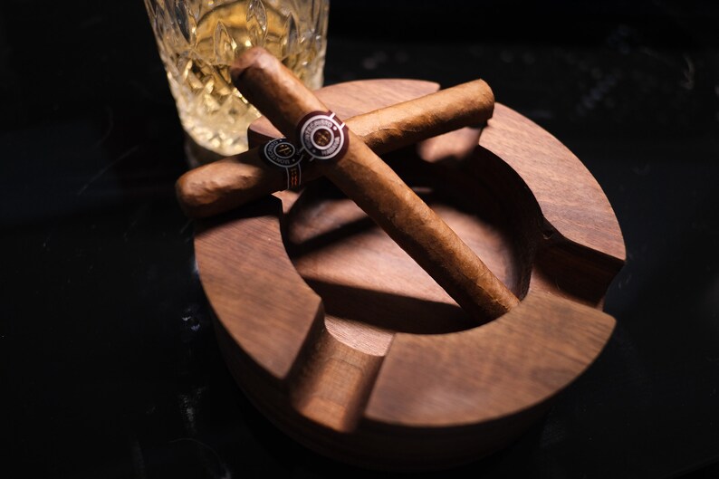 May include: A wooden cigar ashtray with two brown cigars resting inside. The cigars are crossed over each other and have a gold band with a red and white logo.