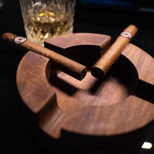 May include: Two brown cigars resting in a wooden cigar ashtray. The ashtray is made of dark wood and has a round shape.