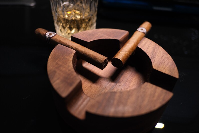 May include: Two brown cigars resting in a wooden cigar ashtray. The ashtray is made of dark wood and has a round shape.