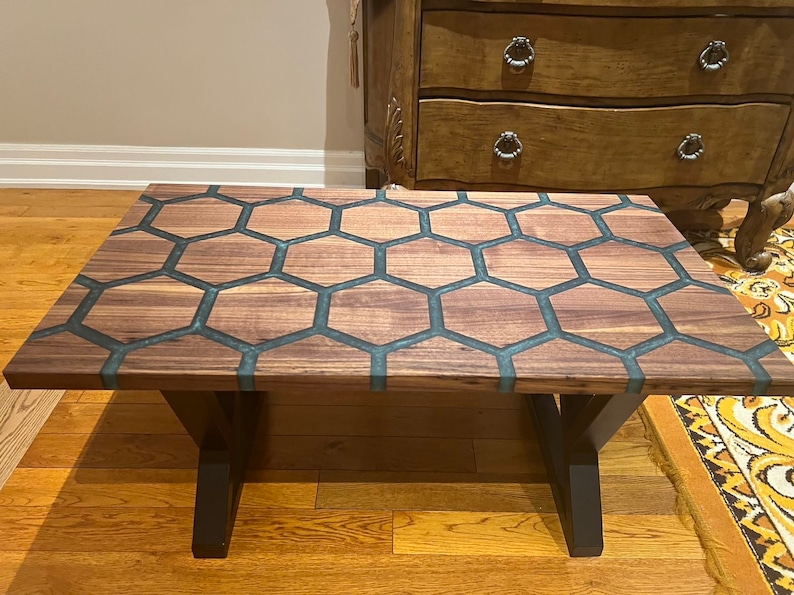 May include: A rectangular coffee table with a unique honeycomb design. The tabletop features a pattern of wooden hexagons filled with a teal-colored resin. The table has dark brown legs and a natural wood grain finish.
