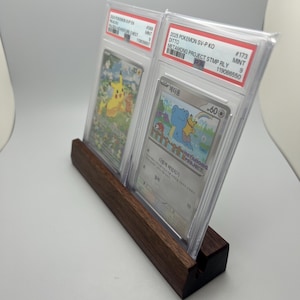 May include: A wooden display stand holding two graded Pok&eacute;mon trading cards. The cards are encased in protective cases, featuring colorful illustrations of Pok&eacute;mon characters. The stand is made of dark wood and has a slot to hold the cards upright.