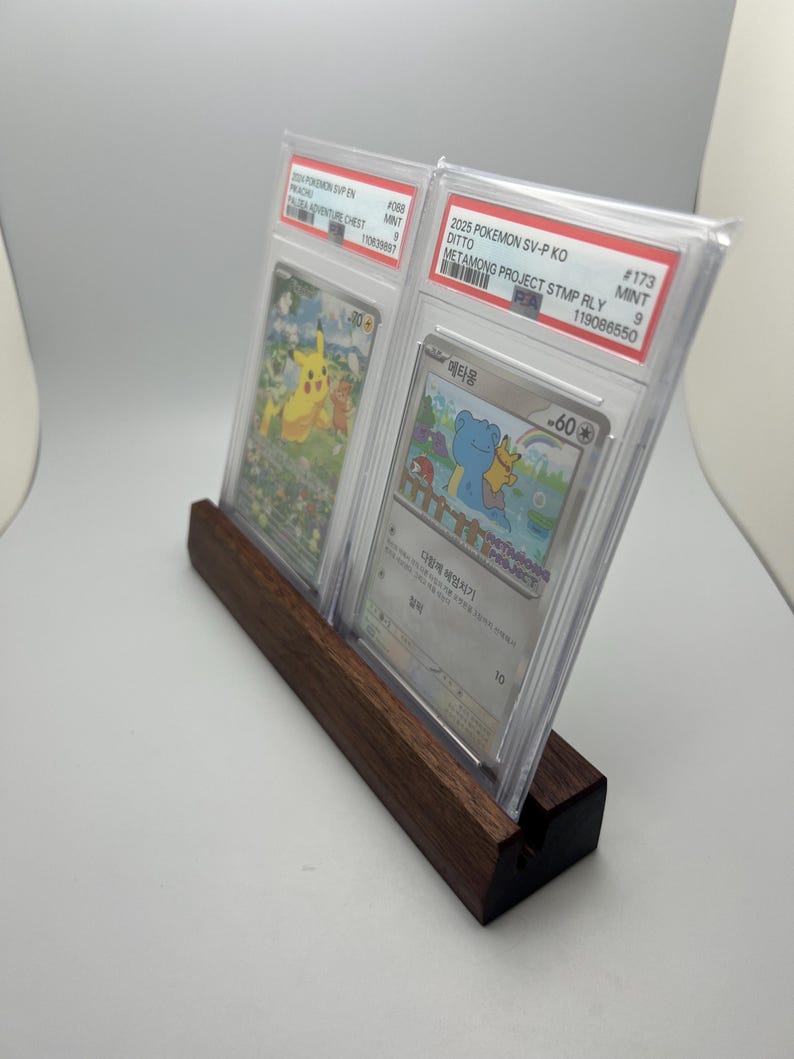 May include: A wooden display stand holding two graded Pok&eacute;mon trading cards. The cards are encased in protective cases, featuring colorful illustrations of Pok&eacute;mon characters. The stand is made of dark wood and has a slot to hold the cards upright.