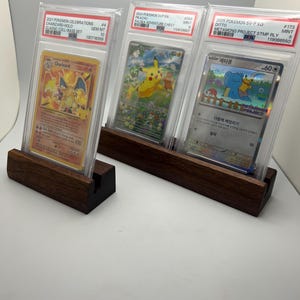 May include: Three graded Pok&eacute;mon trading cards displayed in clear cases, each resting on a dark wooden stand. The cards feature Charizard, Pikachu, and Ditto, with text indicating the set and grading details. The stands are rectangular with a slot to hold the cards.