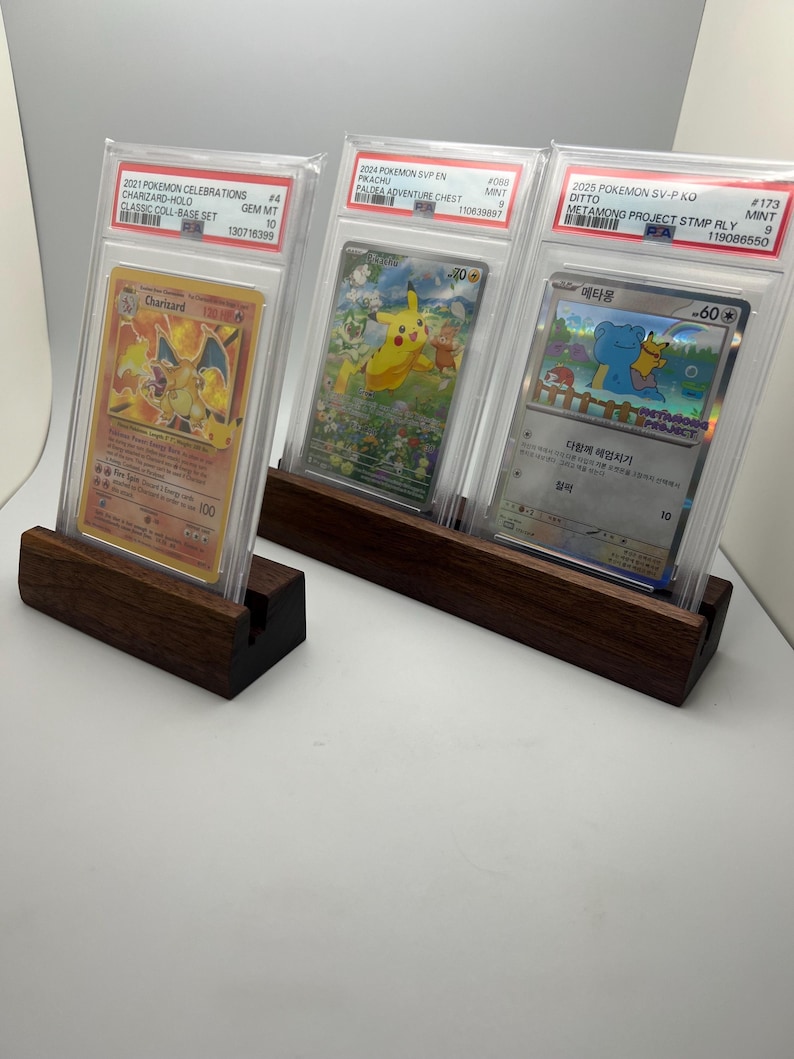 May include: Three graded Pok&eacute;mon trading cards displayed in clear cases, each resting on a dark wooden stand. The cards feature Charizard, Pikachu, and Ditto, with text indicating the set and grading details. The stands are rectangular with a slot to hold the cards.