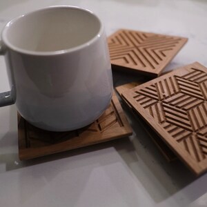 May include: A white ceramic mug with a gray handle rests on a wooden coaster. Three additional wooden coasters with geometric patterns are visible. The coasters are square and made of light brown wood.