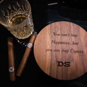 May include: A wooden coaster with the engraved text "You can't buy Happiness, but you can buy Cigars" and the initials "DS". A glass of whiskey and two cigars are on a black surface next to the coaster.