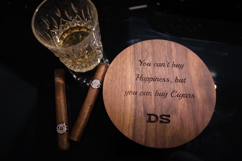 May include: A wooden coaster with the engraved text "You can't buy Happiness, but you can buy Cigars" and the initials "DS". A glass of whiskey and two cigars are on a black surface next to the coaster.
