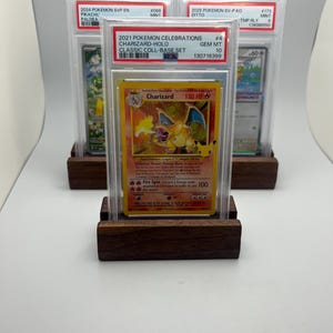 May include: Three graded Pok&eacute;mon trading cards, including a Charizard-Holo card, displayed on wooden stands. The Charizard card is from the 2021 Pok&eacute;mon Celebrations Classic Coll-Base Set. The cards are encased in protective cases.