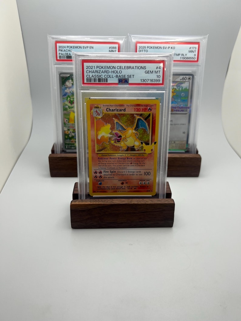May include: Three graded Pok&eacute;mon trading cards, including a Charizard-Holo card, displayed on wooden stands. The Charizard card is from the 2021 Pok&eacute;mon Celebrations Classic Coll-Base Set. The cards are encased in protective cases.