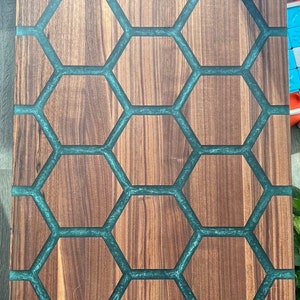 May include: Wooden cutting board with a unique design. The board features a honeycomb pattern with teal-colored resin filling the spaces between the wooden hexagons. The wood has a natural grain, and the board is rectangular.