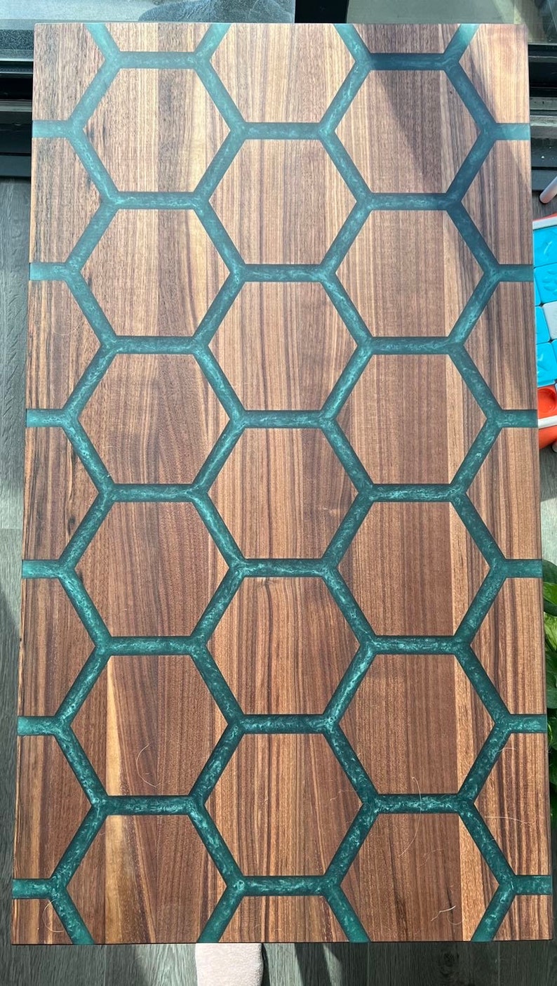 May include: Wooden cutting board with a unique design. The board features a honeycomb pattern with teal-colored resin filling the spaces between the wooden hexagons. The wood has a natural grain, and the board is rectangular.