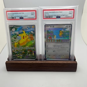 May include: Two Pokemon trading cards in protective cases, displayed on a dark wooden stand. The cards feature Pikachu and Ditto, with text including "2024 Pokemon SVP EN" and "2025 Pokemon SV-P KO". The Pikachu card has a value of 70.
